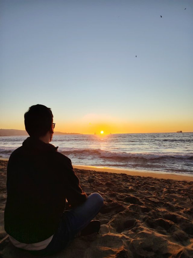 Cristóbal Sanhueza Rodríguez sentado en la playa, de espaldas, mirando el atardecer sobre el mar
